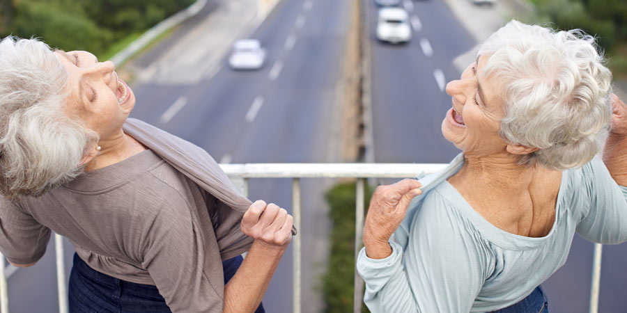 Two women standing on an overpass laughing and lifting their tops to flash the cars below