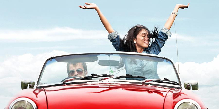 Couple driving a bright red vintage convertible and the woman has her hands in the air 