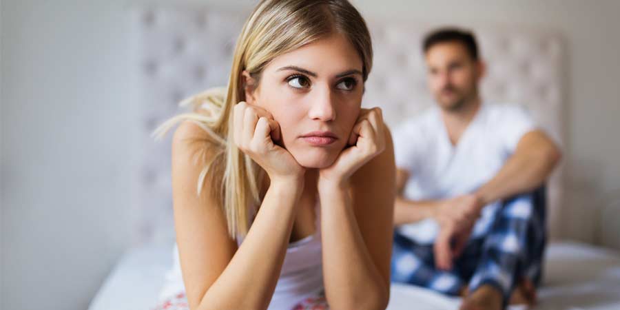 Woman sitting on edge of bed in thought while her partner sits in the background