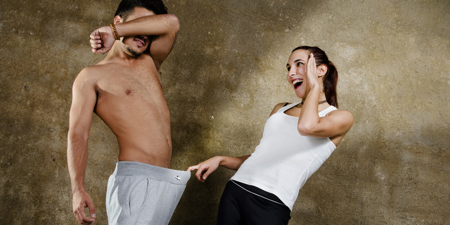 Woman looking down a man's tracksuit pants and laughing enviously at his penis