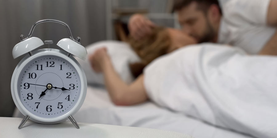 Couple in bed in the background with an alarm clock in the foreground showing the time